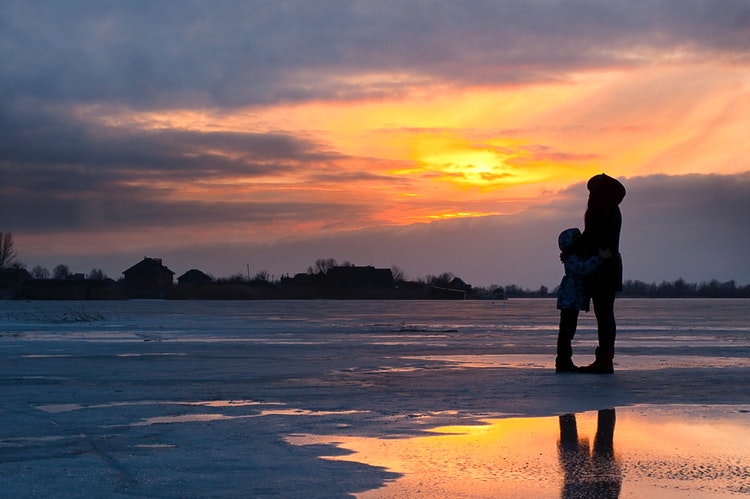 Mother and child standing on the shore of a snowy beach at sunset
